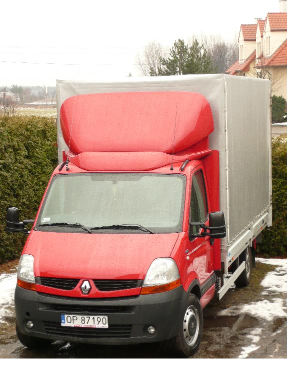 Czerwony samochód dostawczy Renault z plandeką w pochmurny dzień, widoczny numer rejestracyjny OP 87190, zarys zabudowań mieszkalnych w tle.